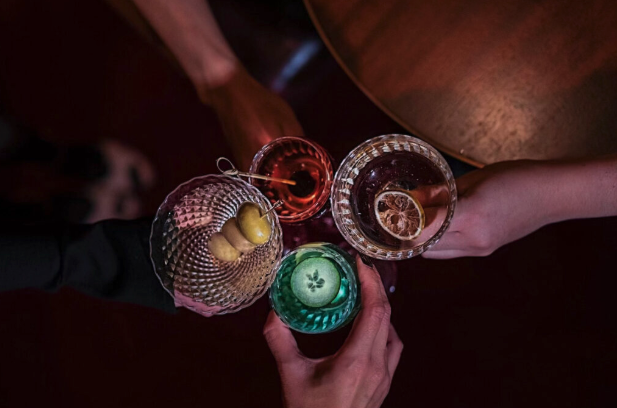 Four hands holding assorted cocktails with garnishes including olives, cucumber, and citrus, raised in a toast above a dimly lit bar table.
