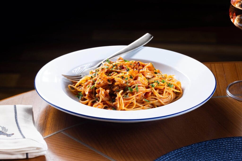 Restaurants at Encore Boston Harbor Resort Close-up of a white plate with spaghetti in a tomato-based sauce garnished with herbs and parmesan, served on a wooden table with a cloth napkin and glass nearby.