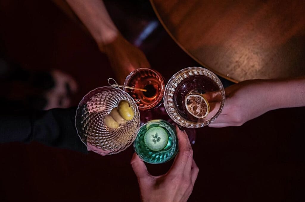 Overhead view of four hands toasting with various cocktails in cut-glass cups, each garnished uniquely with olives, lemon, or cucumber.